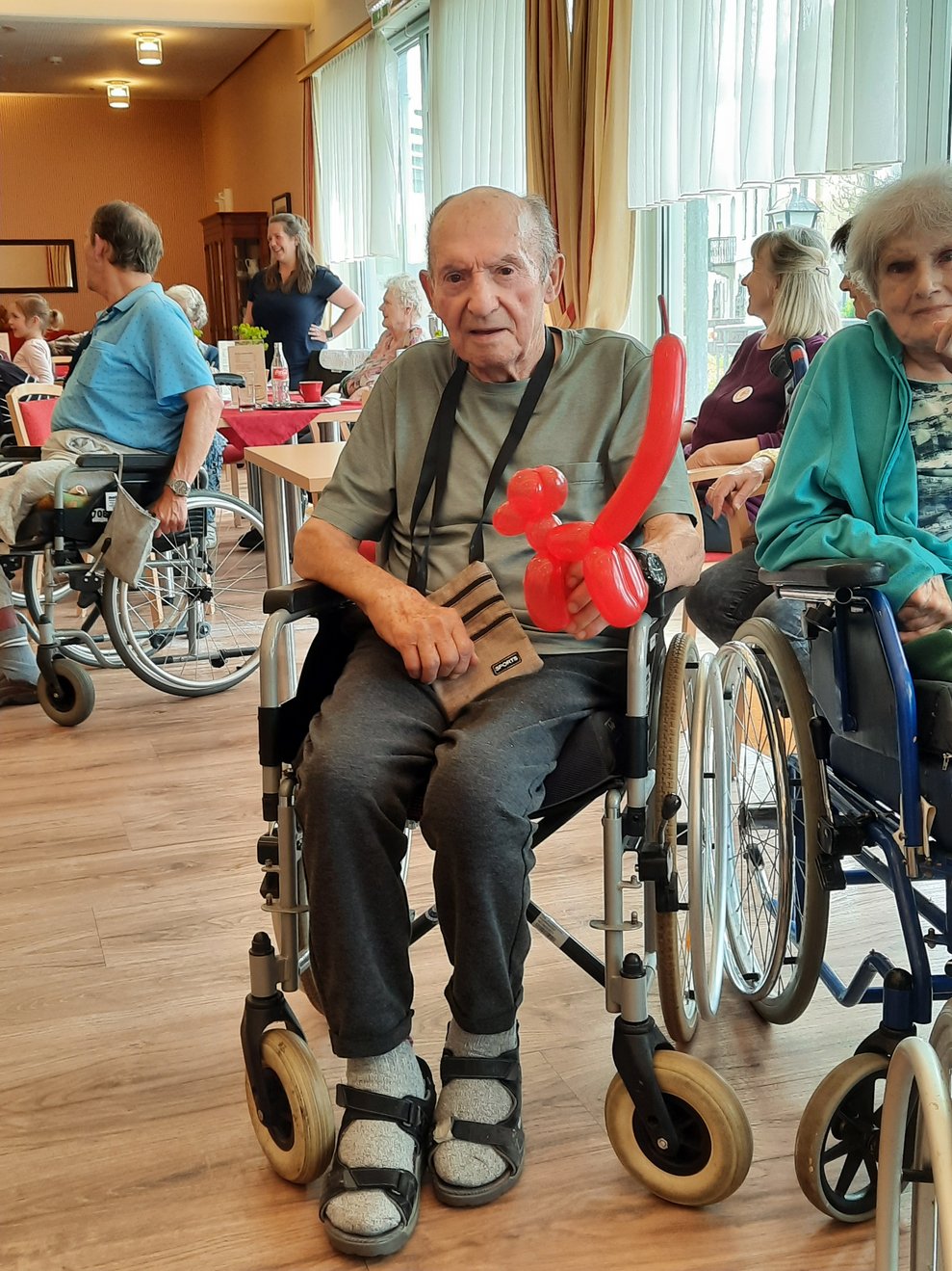 Ein Herr im Rollstuhl hält ein rotes Lufballontier in die Kamera.