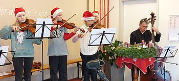 Vier Kinder musizieren mit Streichinstrumenten und haben Weihnachtsmützen am Kopf.